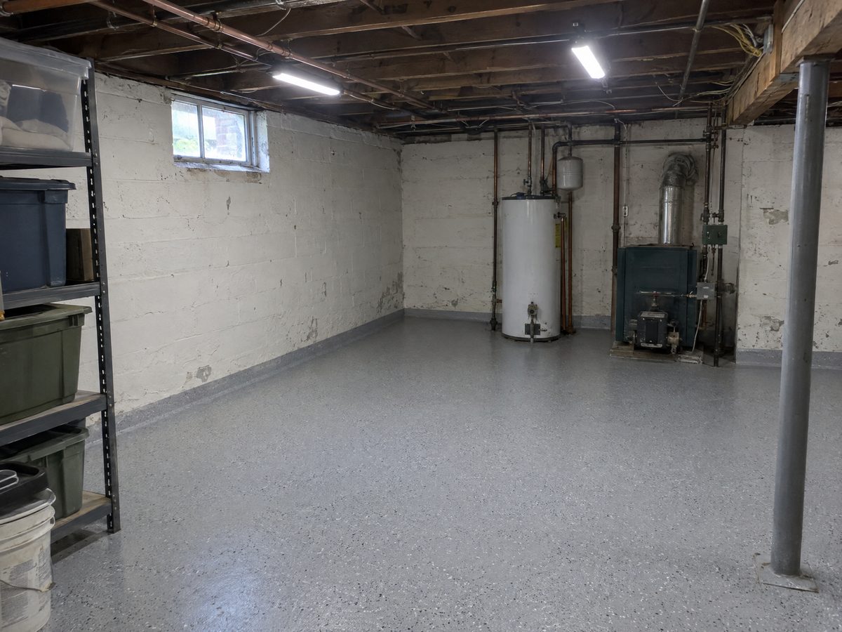 A basement with painted block walls, a water heater and an oil burner, and a new light grey flake floor coating with a short cove at the walls