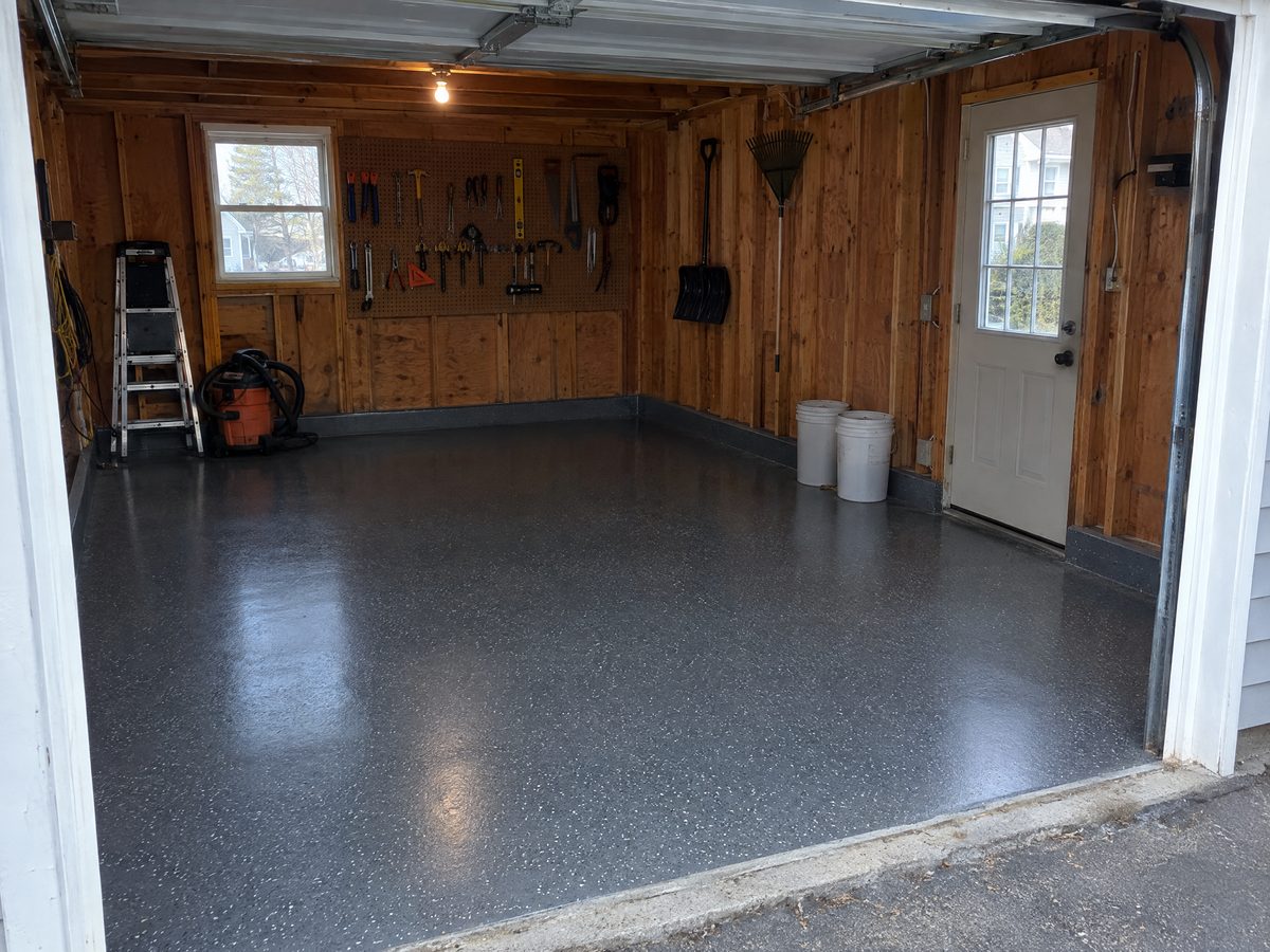 A single-bay wooden garage with a charcoal flake floor coating turned up the walls as a cove, tools on a pegboard and two empty buckets by the side door