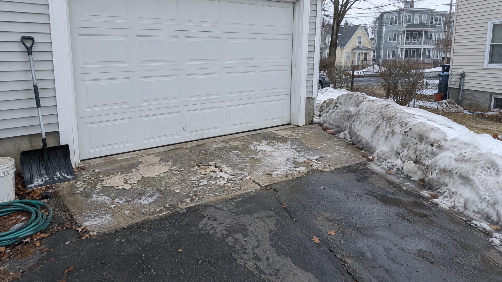 A garage apron in late winter, its concrete scaled and flaking with salt crust in the low spots, beside a plowed snowbank and a wet driveway