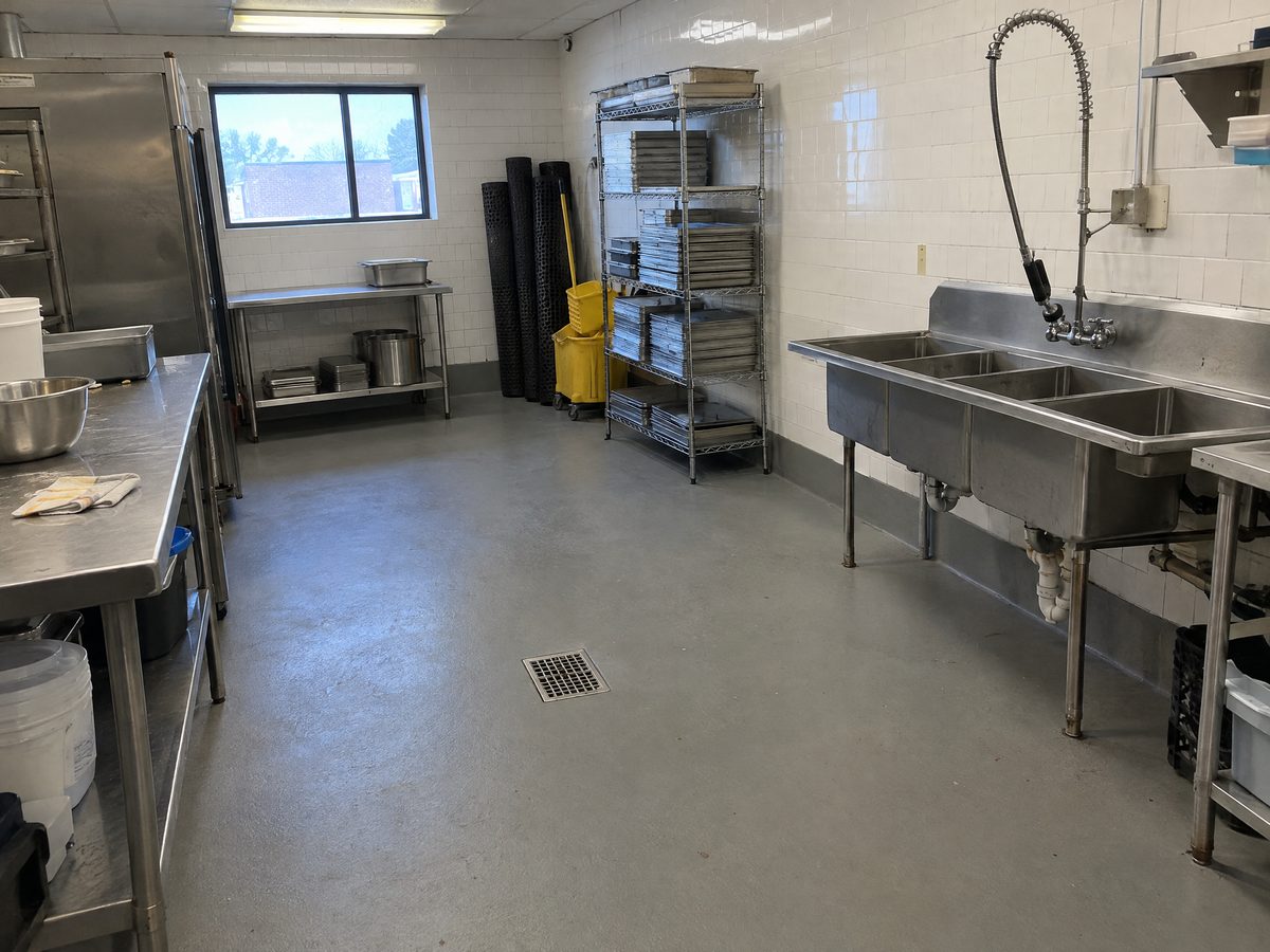 A restaurant back kitchen with a matte grey urethane cement floor, a coved base along the tile wall, a floor drain and stainless prep tables