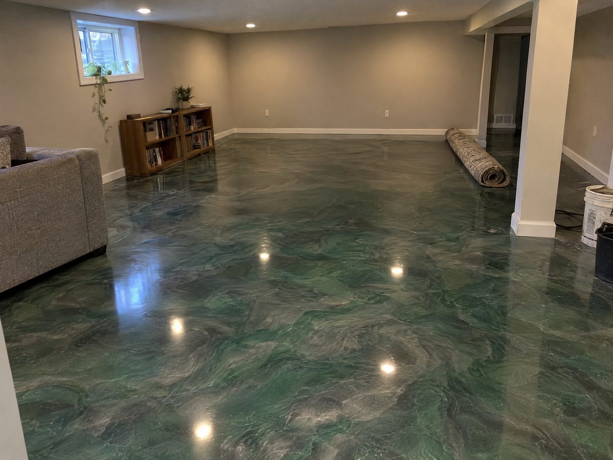 A basement family room with a green and pewter metallic epoxy floor, a grey sofa, a wooden bookcase and a rolled rug waiting to go down